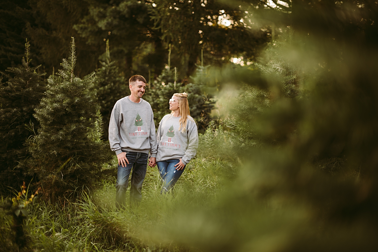 A couple wearing matching gray Christmas sweatshirts stands together, holding hands and smiling at each other during their engagement session at Middleton Tree Farm, surrounded by lush green pine trees.
