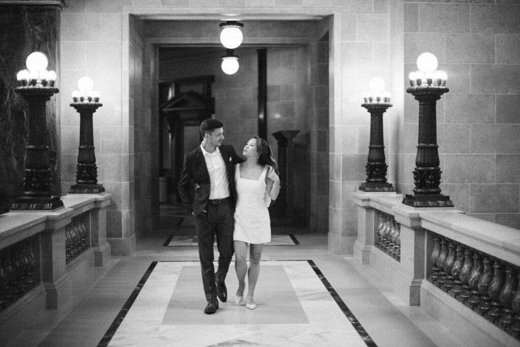 A couple dressed formally walks arm-in-arm down a grand hallway with ornate lighting and marble walls, smiling at each other. The black-and-white image exudes classic elegance—perfect inspiration for those exploring Wisconsin photographer pricing.