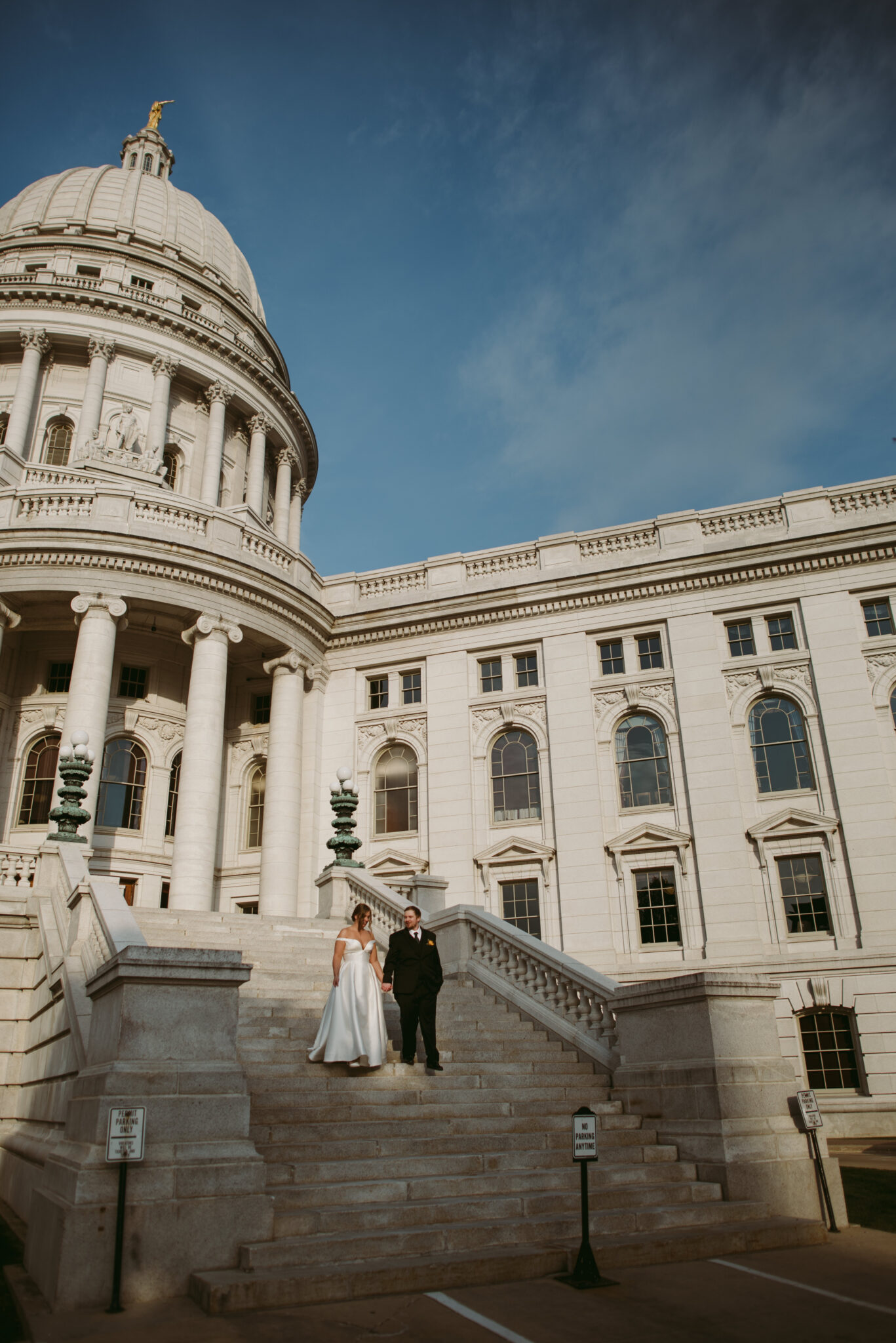 A newlywed couple stands on the steps of the Wisconsin State Capitol under a blue sky. The bride in white and groom in black hold hands on the stone staircase, capturing a perfect elopement moment outside this grand, white-domed building.