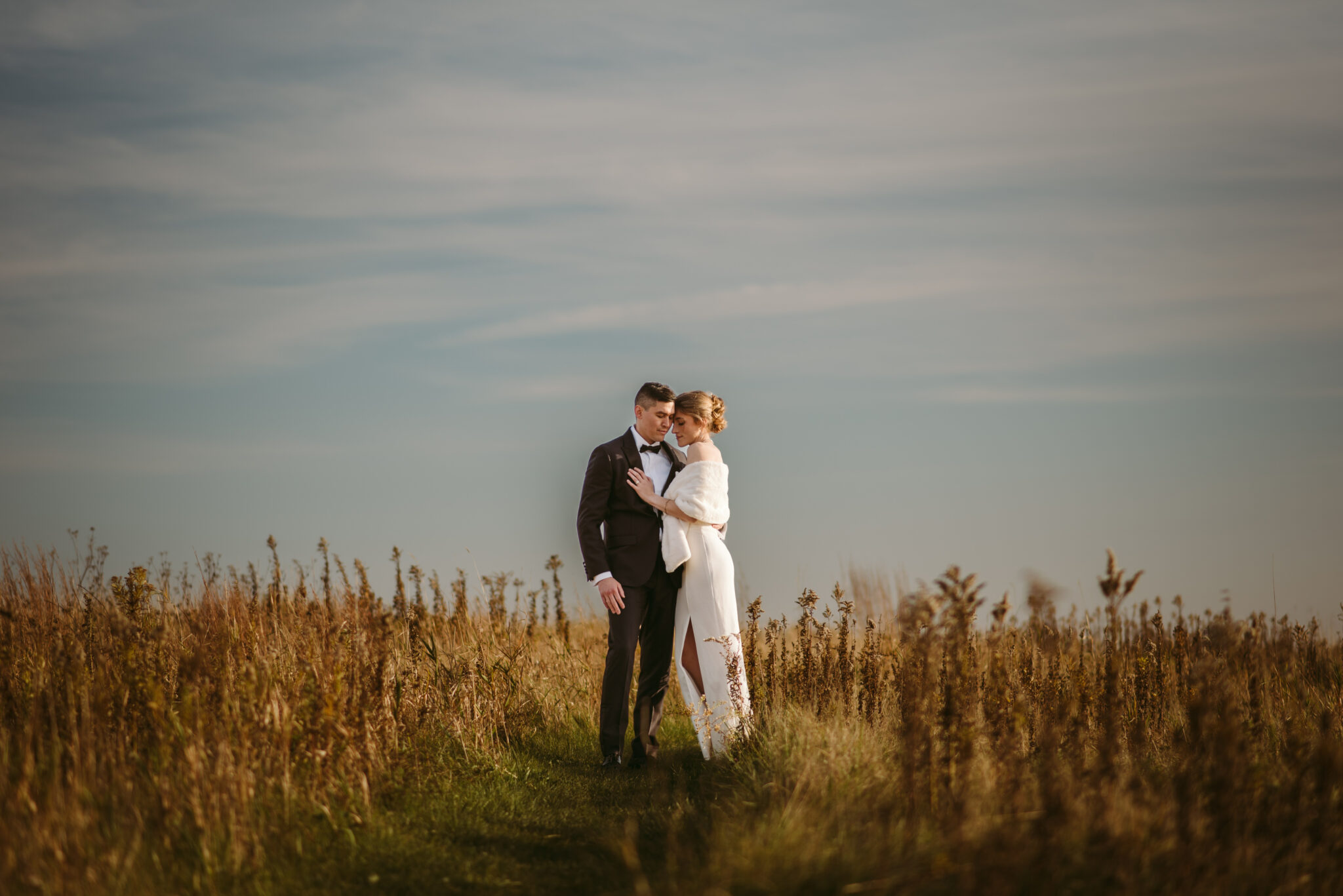 A bride and groom stand close together in a grassy field under a blue sky with wispy clouds, sharing an intimate moment surrounded by tall grass on the day they chose to elope in 6 weeks.