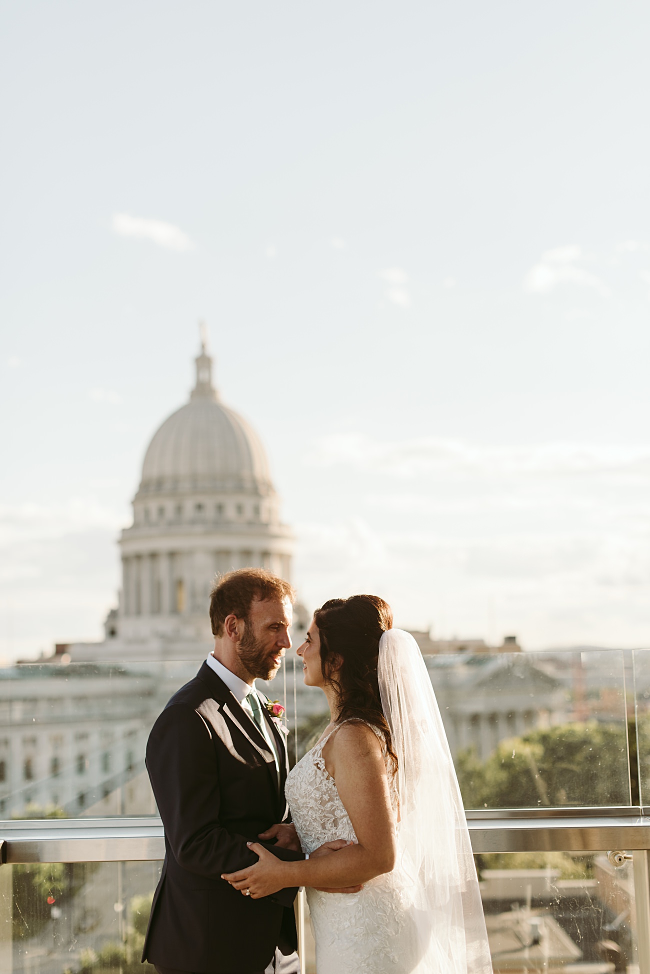 A bride and groom stand close together on a rooftop, gazing at each other, with the state capitol wedding venue’s domed building visible in the background under a bright sky.