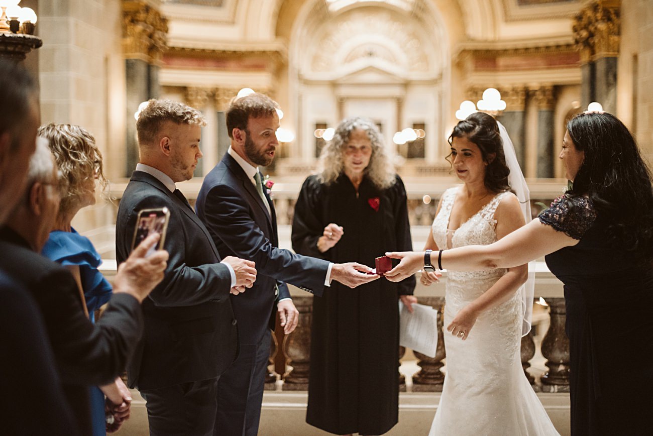 A bride and groom exchange rings during a state capitol wedding ceremony, surrounded by guests and an officiant in an ornate indoor setting with arches and elegant lighting.