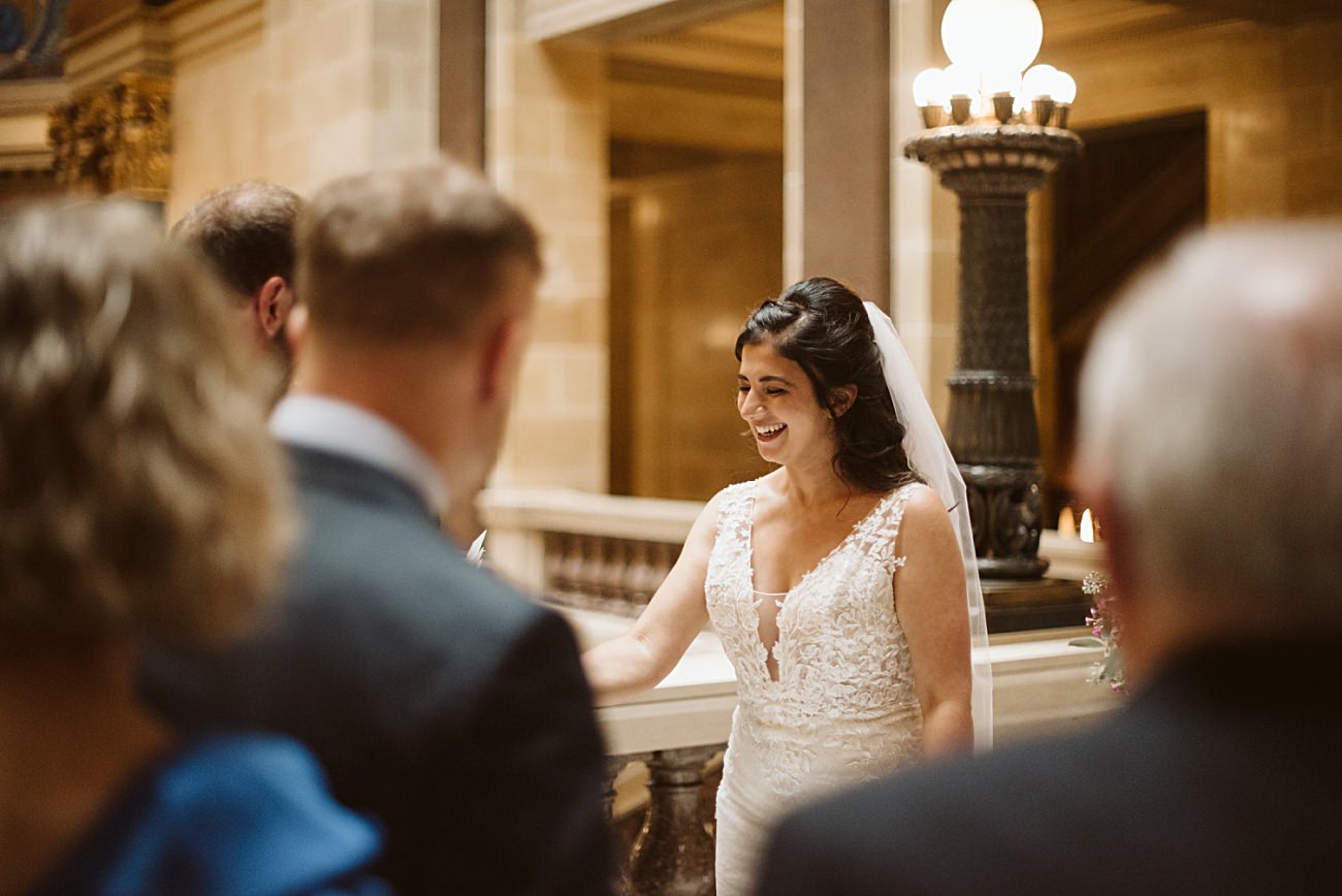 A bride in a lace wedding dress and veil smiles warmly while standing indoors at a state capitol wedding, facing a small group of people in formal attire. Elegant lighting and grand stone architecture enhance the ceremony’s sophisticated setting.