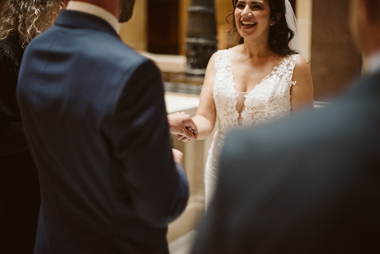 A bride in a lace wedding dress smiles and holds hands with a groom in a blue suit during their state capitol wedding ceremony indoors. The image captures a joyful, candid moment between the couple.