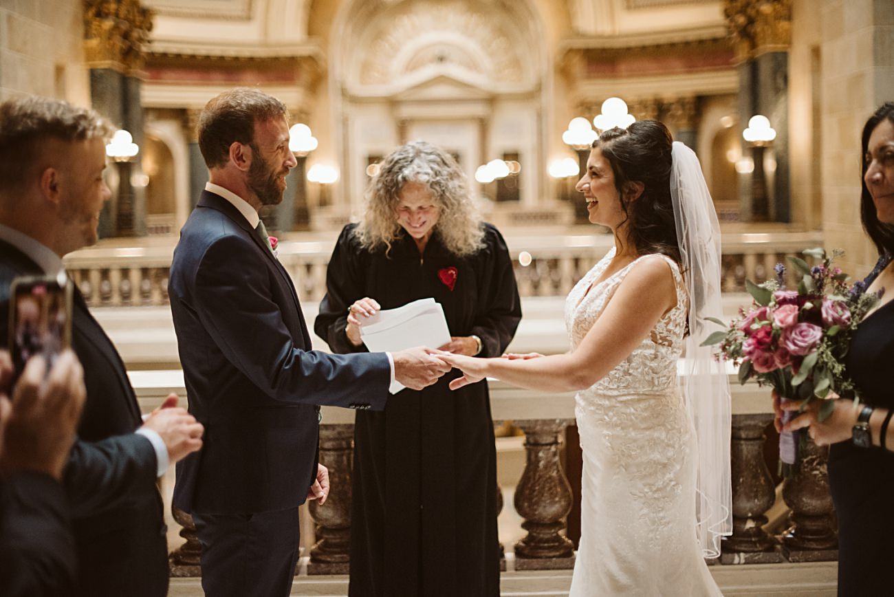 A bride and groom exchange rings during a state capitol wedding at an ornate indoor venue, with an officiant behind them and guests nearby. The bride holds a bouquet and wears a veil, while the groom is in a blue suit.