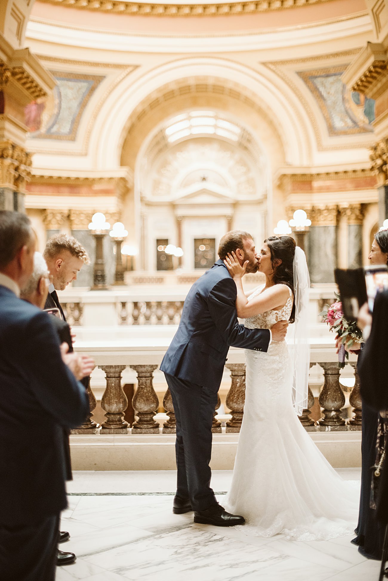 A bride and groom share a kiss during their state capitol wedding in an ornate, marble-floored hall with arched ceilings, while guests stand nearby, capturing the moment.
