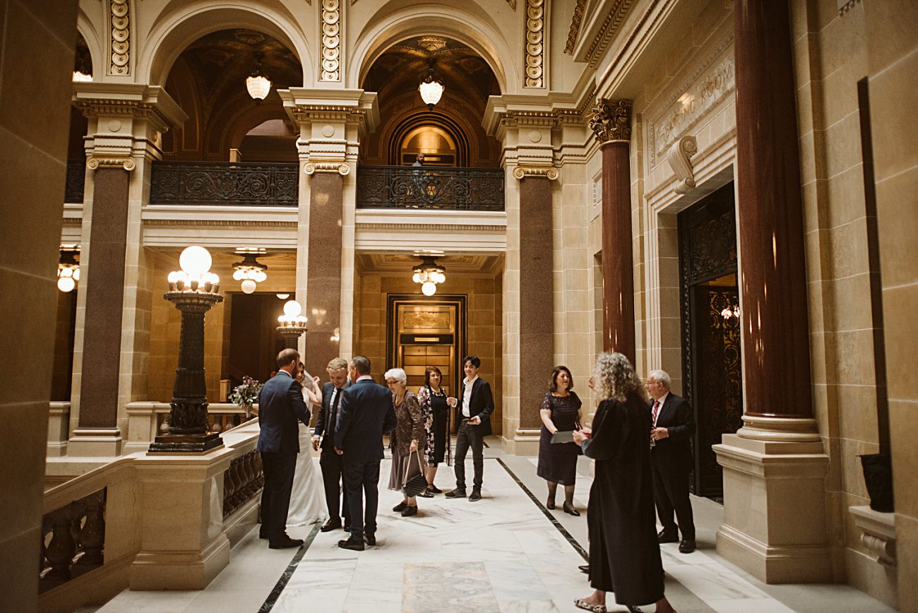 A group of people dressed formally are gathered and conversing in an ornate, marble-floored hall with tall columns, elegant lighting, and arched ceilings—a stunning venue for a state capitol wedding or special event.