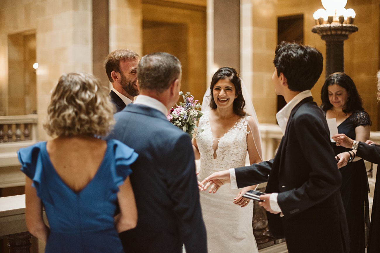 A bride in a white dress smiles joyfully while holding a bouquet, surrounded by well-dressed guests inside an elegant, warmly lit state capitol wedding venue with marble columns and ornate lamps.
