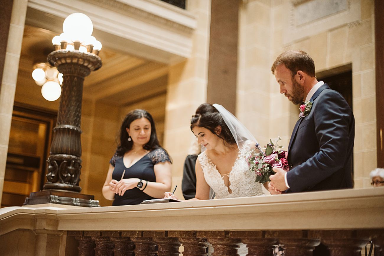 A bride in a white dress and veil signs a document on a stone railing at a state capitol wedding, holding a bouquet, while a groom in a suit and a woman in a navy dress stand beside her in an elegant, warmly lit interior.