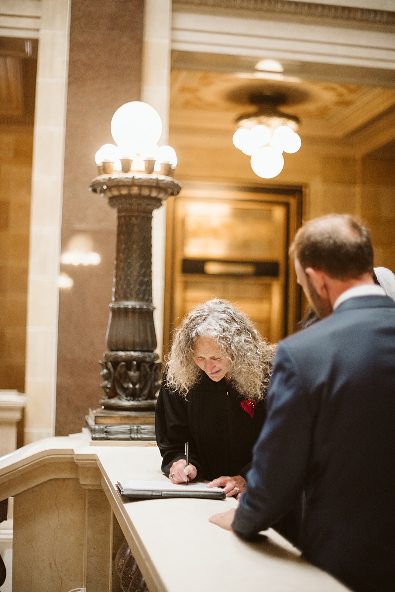 A woman with curly gray hair signs a document at a marble counter in an ornate state capitol building, suggesting a wedding ceremony, while a man in a suit stands nearby amid decorative columns and warm lighting.