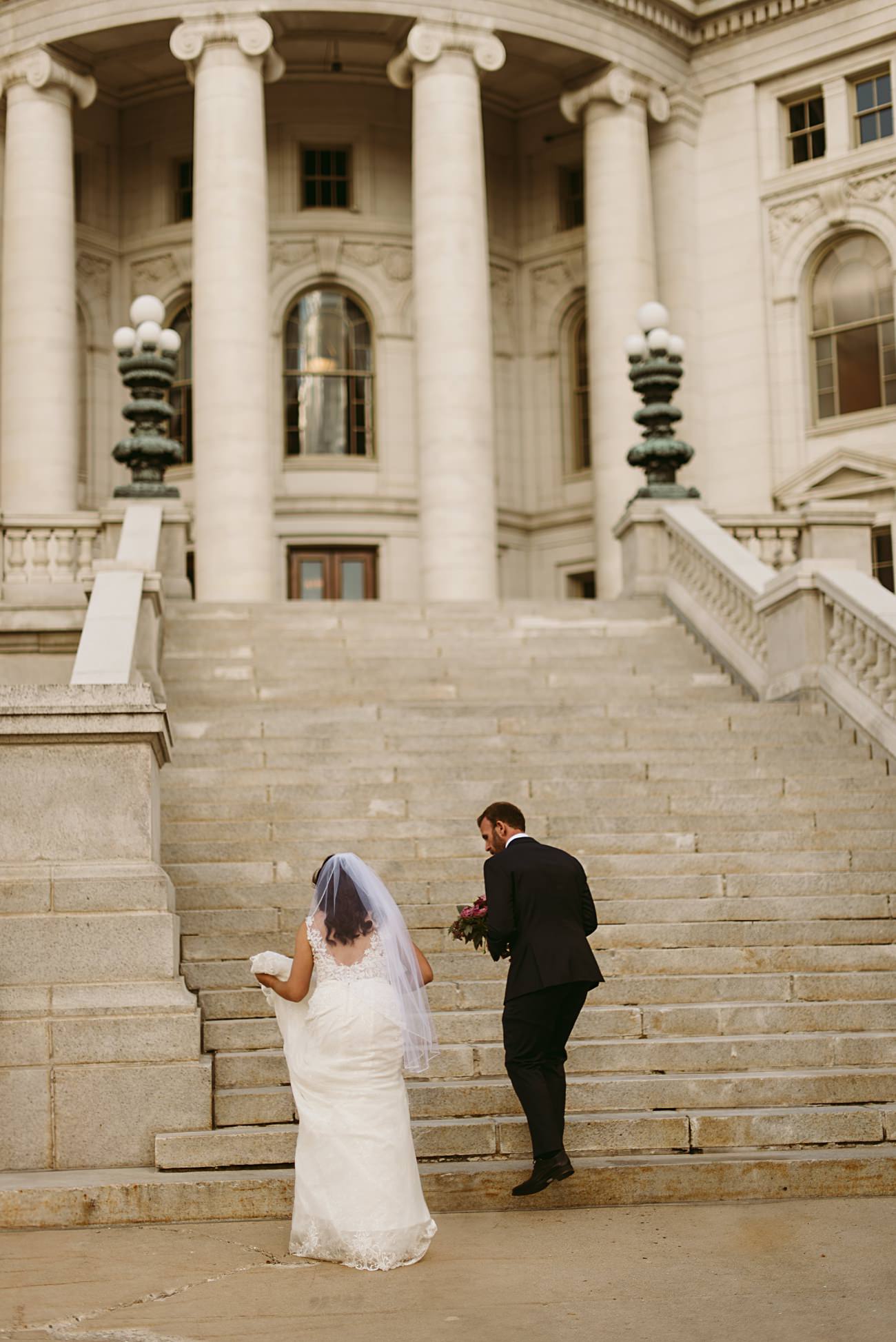 A bride in a white dress and veil walks up stone steps beside a groom in a black suit, leading to a grand state capitol building with tall columns and ornate details, creating an elegant wedding scene.