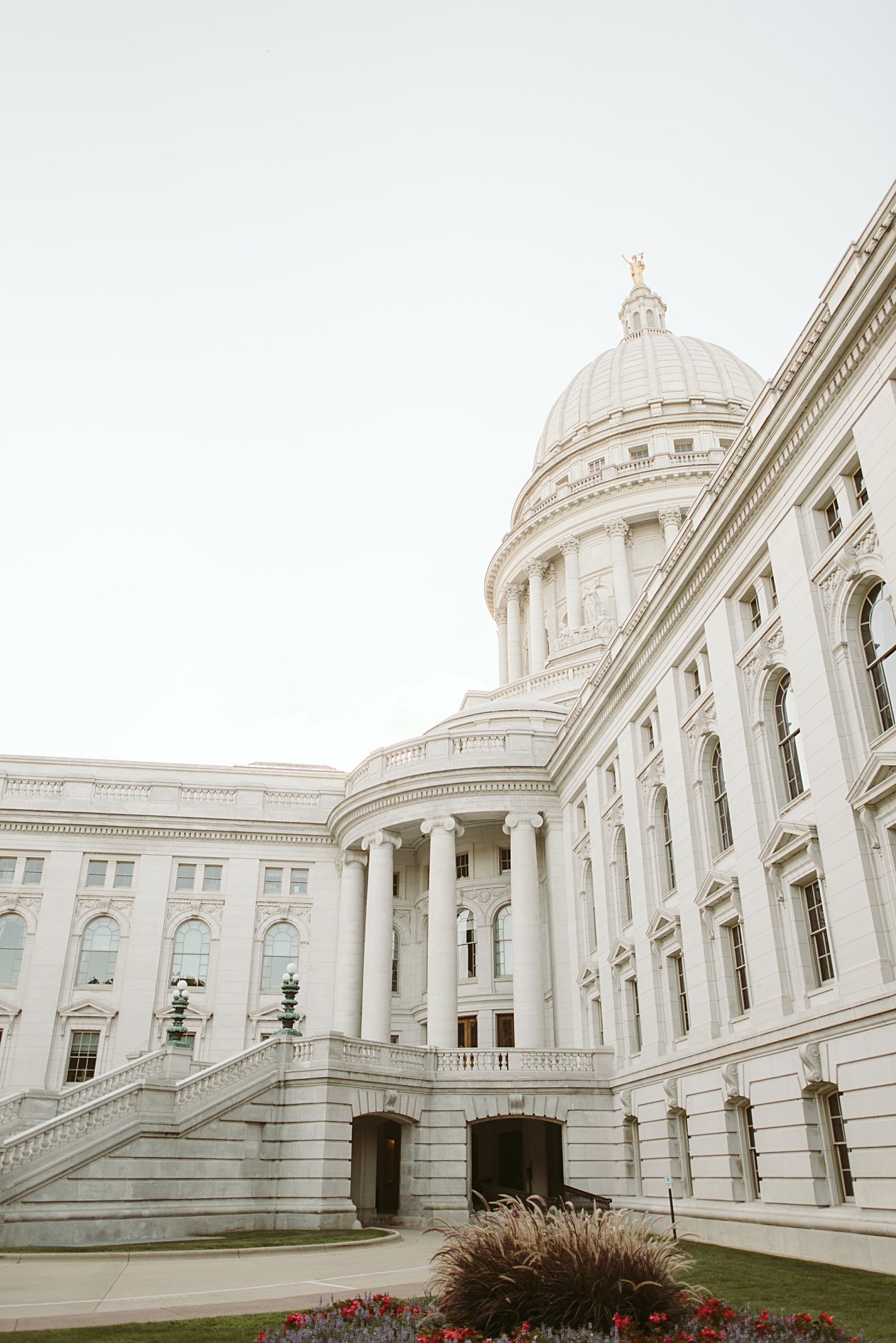 A grand white stone state capitol building with a large dome, columns, and arched windows is photographed from the courtyard. The clear sky and blooming flowers in the foreground create a picturesque setting for a stunning state capitol wedding.