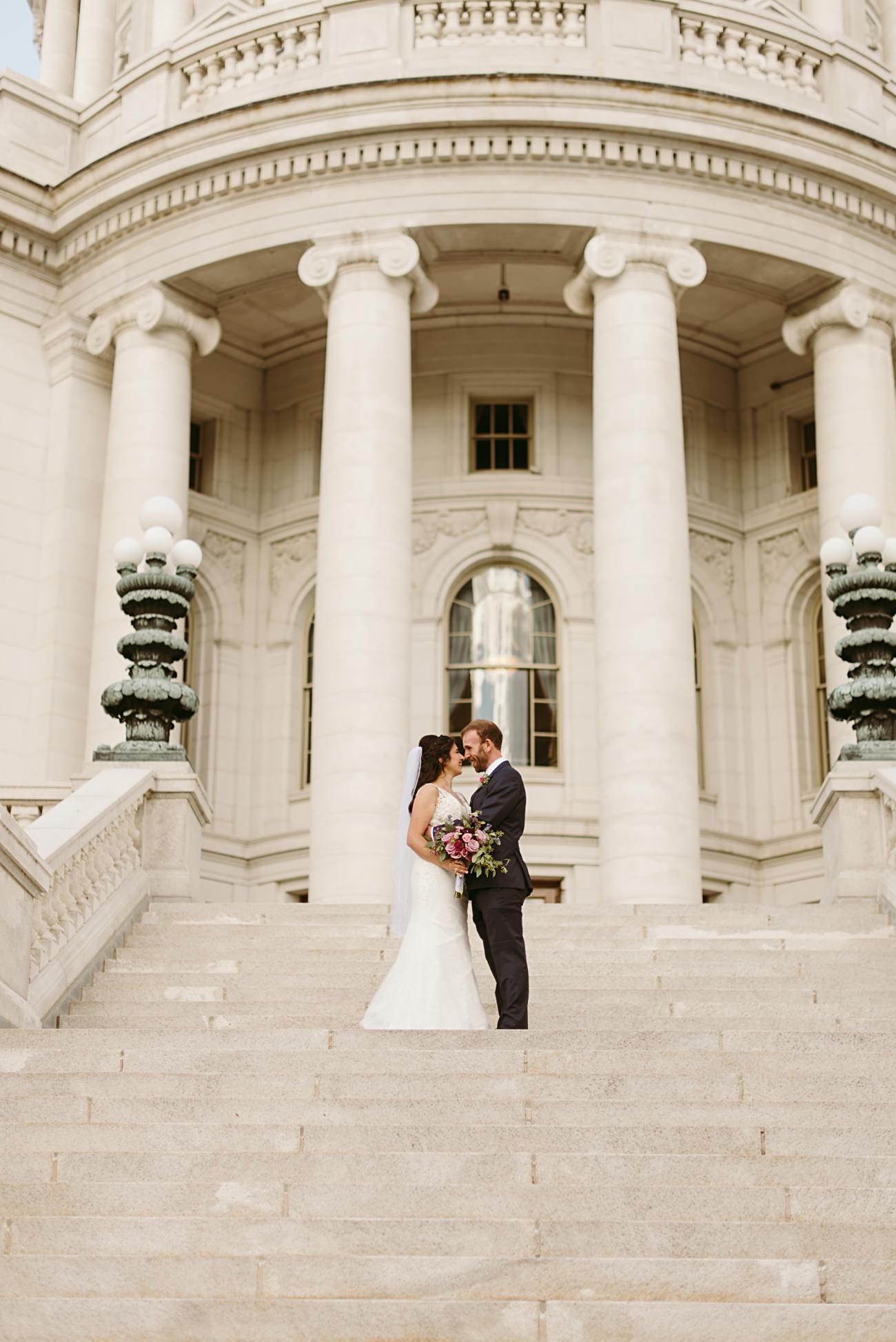 A bride and groom stand close together on stone steps, holding a bouquet and facing each other, in front of a grand state capitol building with tall columns and ornate lamps—capturing an elegant wedding moment.