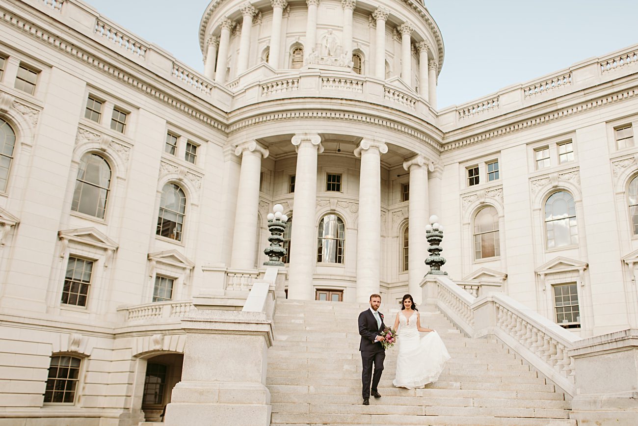 A bride and groom walk down the steps of a grand white stone state capitol building with columns and large windows, holding hands and smiling, with the bride holding her dress and the groom holding a bouquet after their elegant wedding.