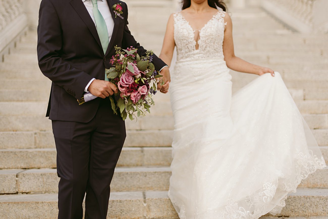 A bride in a white lace wedding gown holds her dress while walking down steps at a state capitol wedding, beside a groom in a dark suit holding a bouquet of purple and pink flowers. Their faces are not visible.