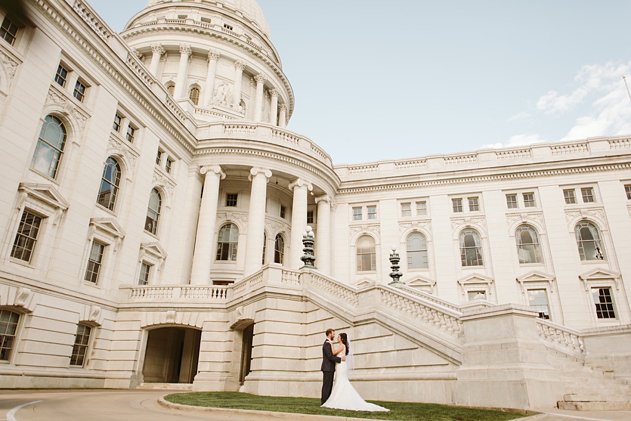 A bride and groom embrace on the steps of a grand, white stone state capitol building with columns and a large dome, likely during their elegant state capitol wedding, under a clear blue sky.