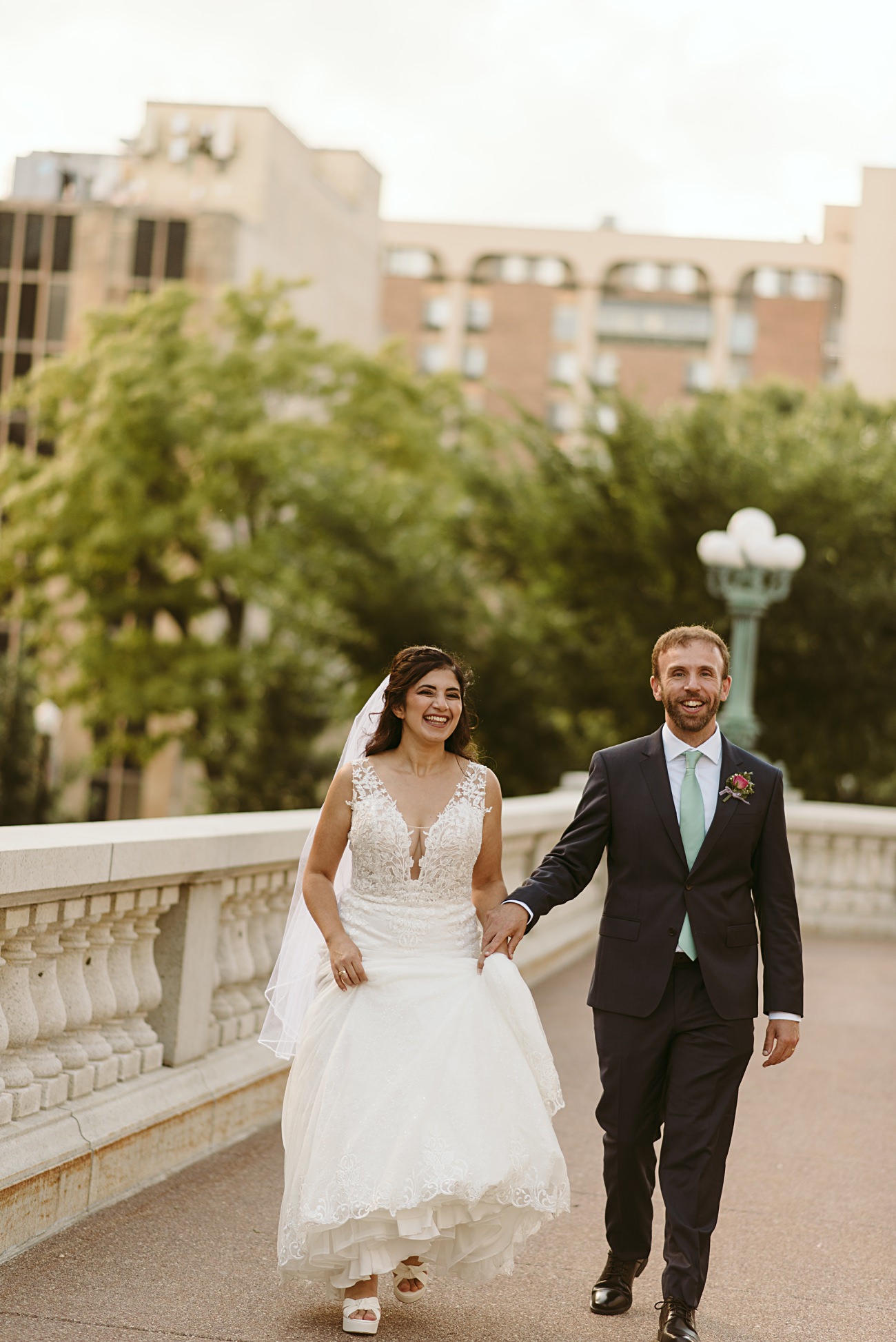 A bride in a white lace dress and a groom in a dark suit with a mint green tie walk hand in hand on a city bridge, both smiling after their state capitol wedding. Trees and buildings are visible in the background.