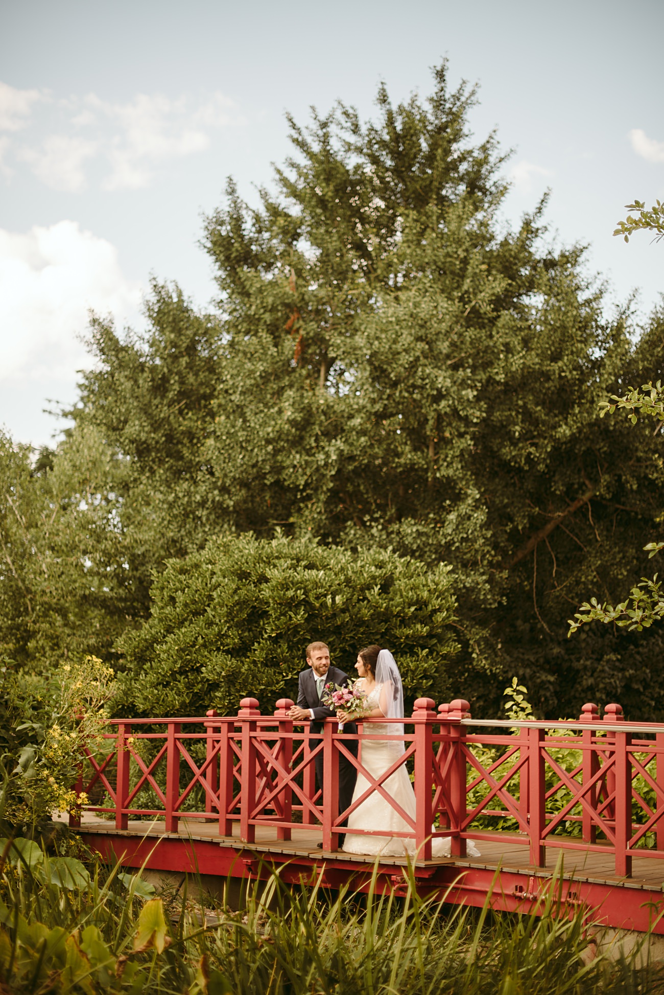 A bride and groom stand facing each other on a red wooden bridge surrounded by lush greenery, sharing a joyful moment on their state capitol wedding day.