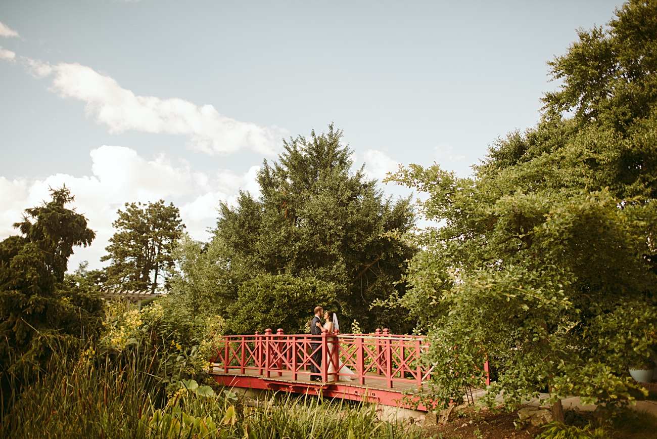 A couple stands together on a red wooden bridge, having just celebrated their state capitol wedding, surrounded by lush green trees and plants under a clear, partly cloudy sky.