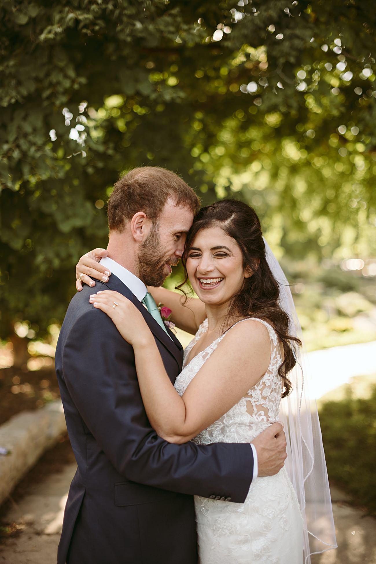 A bride and groom embrace and smile joyfully outdoors under leafy trees near the state capitol wedding venue, with sunlight filtering through the branches. The bride wears a lace wedding dress and veil; the groom wears a dark suit.