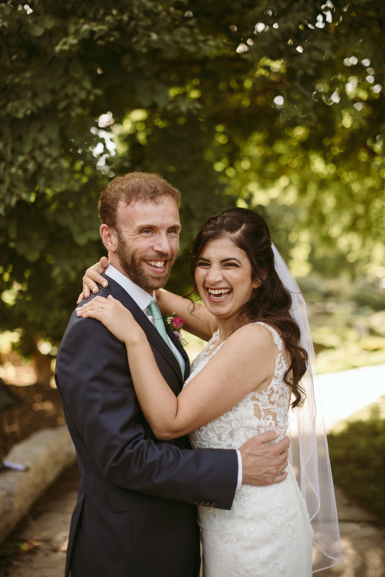 A smiling bride in a white lace dress and veil embraces her groom in a dark suit and green tie at their state capitol wedding, surrounded by greenery and sunlight outdoors.