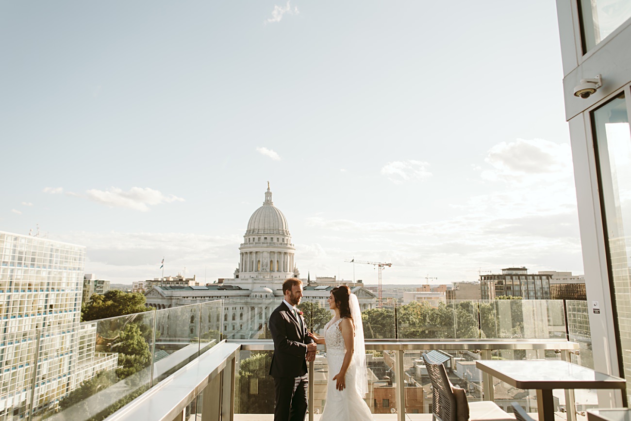 A bride and groom stand on a rooftop terrace, holding hands and facing each other during their state capitol wedding, with a cityscape and the large domed capitol building in the background under a clear sky.