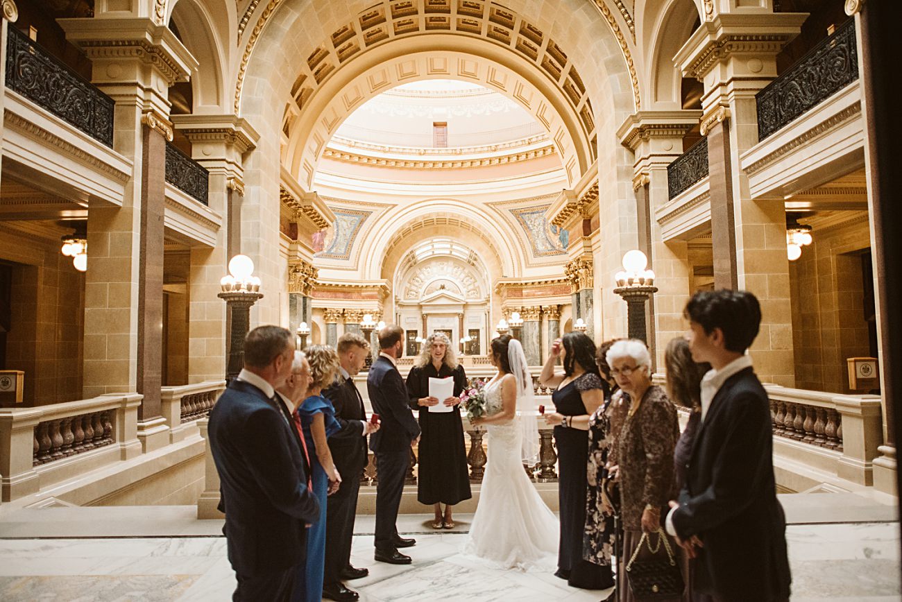 A bride and groom stand before an officiant in an ornate, domed hall at a state capitol wedding, surrounded by family and friends dressed formally, during a ceremony in a grand historic building.