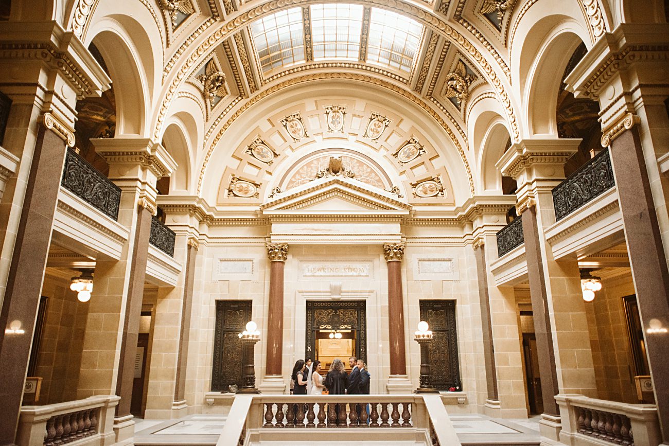 A group of people stands in the grand, ornate lobby of a historic state capitol building, its high arched ceilings and tall columns creating an elegant setting for a wedding celebration beneath large skylights.