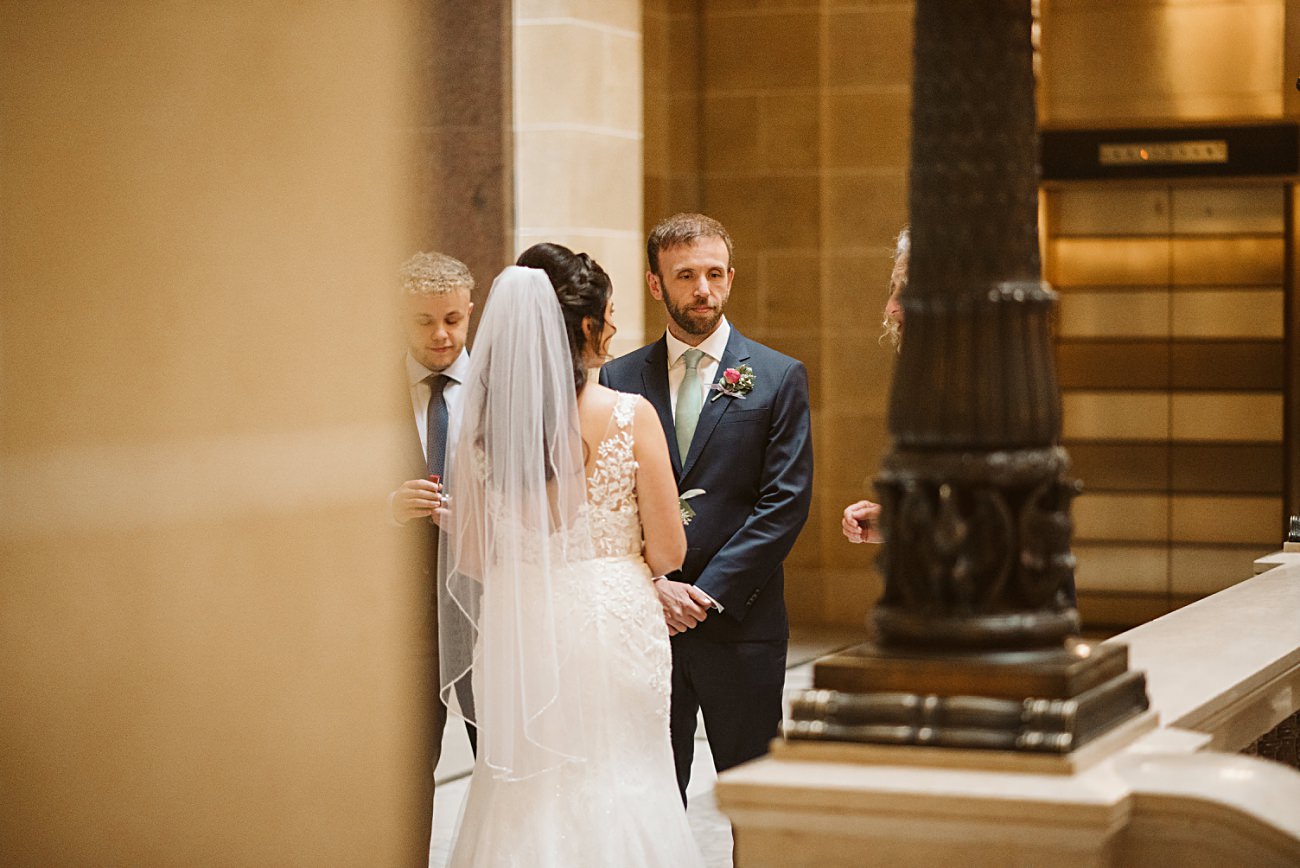 A bride in a white dress and veil stands facing a groom in a suit with a boutonniere indoors at a state capitol wedding, as two others stand nearby in a formal setting with grand stone columns.
