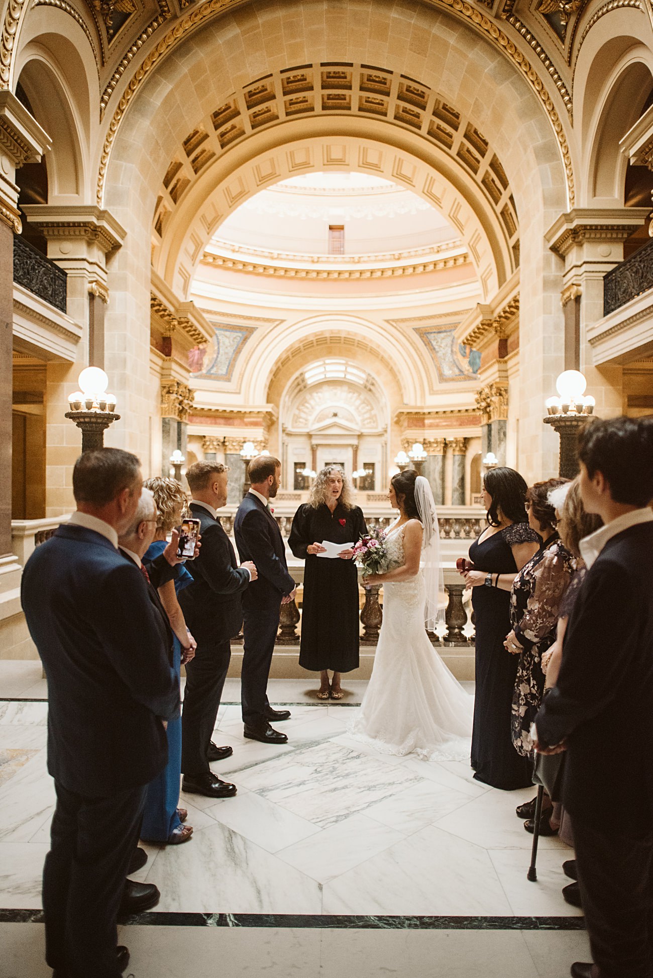 A bride and groom stand together facing an officiant during their state capitol wedding in a grand, ornate hall, surrounded by family and friends. Marble floors and arched ceilings create an elegant atmosphere.