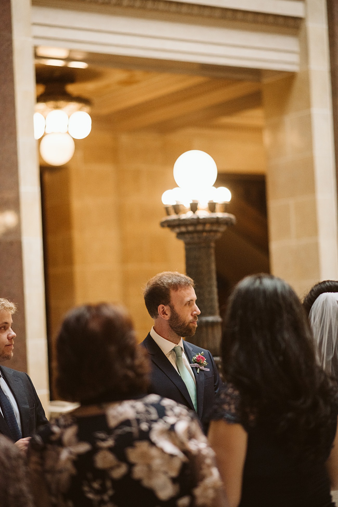 A man in a suit with a green tie and boutonnière stands indoors near a group of people, surrounded by ornate lighting and marble architecture at a state capitol wedding.