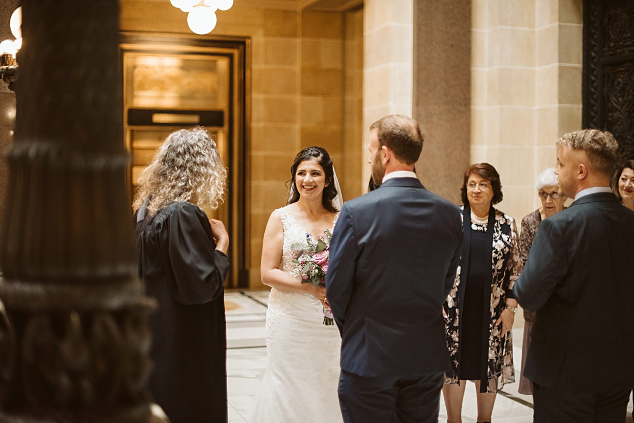 A bride in a white dress and groom in a suit stand together smiling during a state capitol wedding ceremony indoors, surrounded by a small group of people, including an officiant and family members.