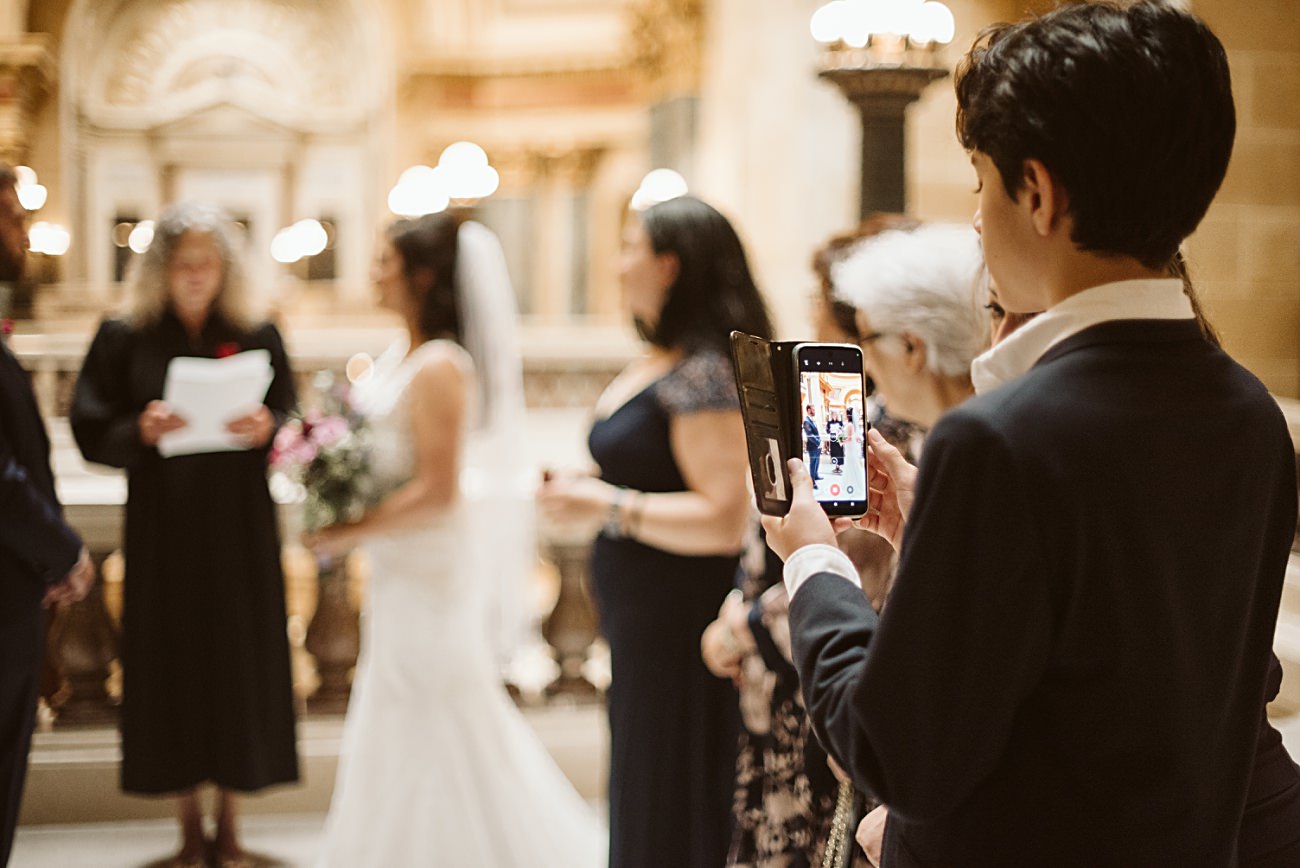 A person records a state capitol wedding on a smartphone while a bride in a white dress and guests stand together indoors, with a judge reading from a paper in the background. The setting appears elegant with warm lighting.